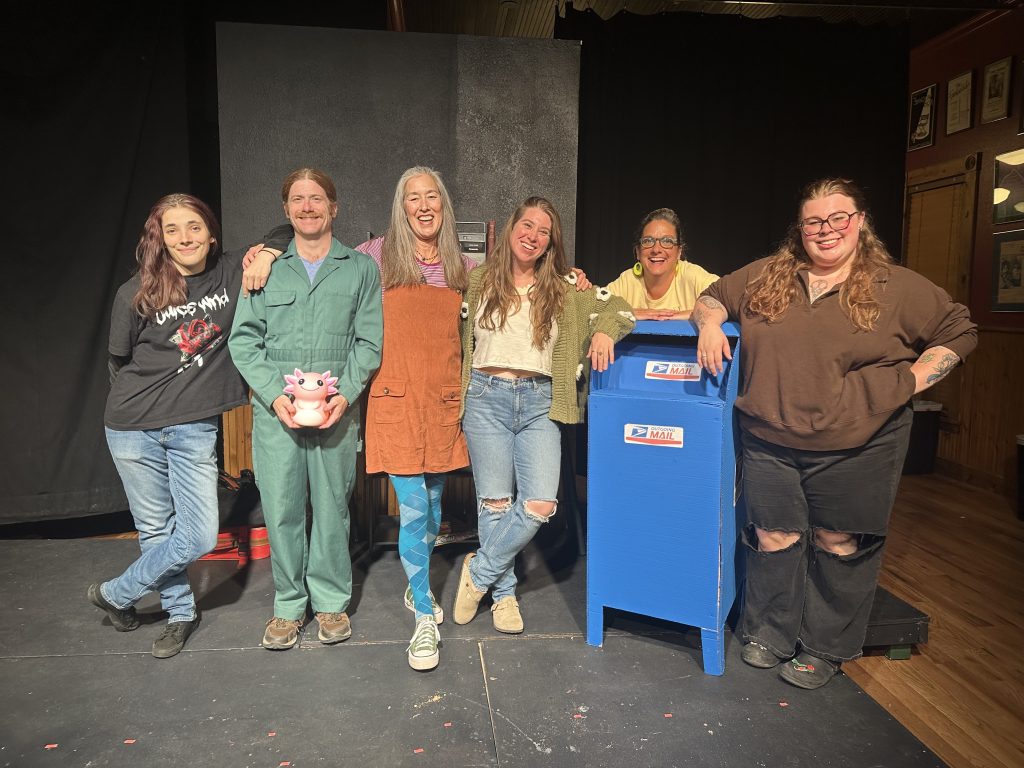 Six cast members stand smiling on stage around a mailbox prop.
