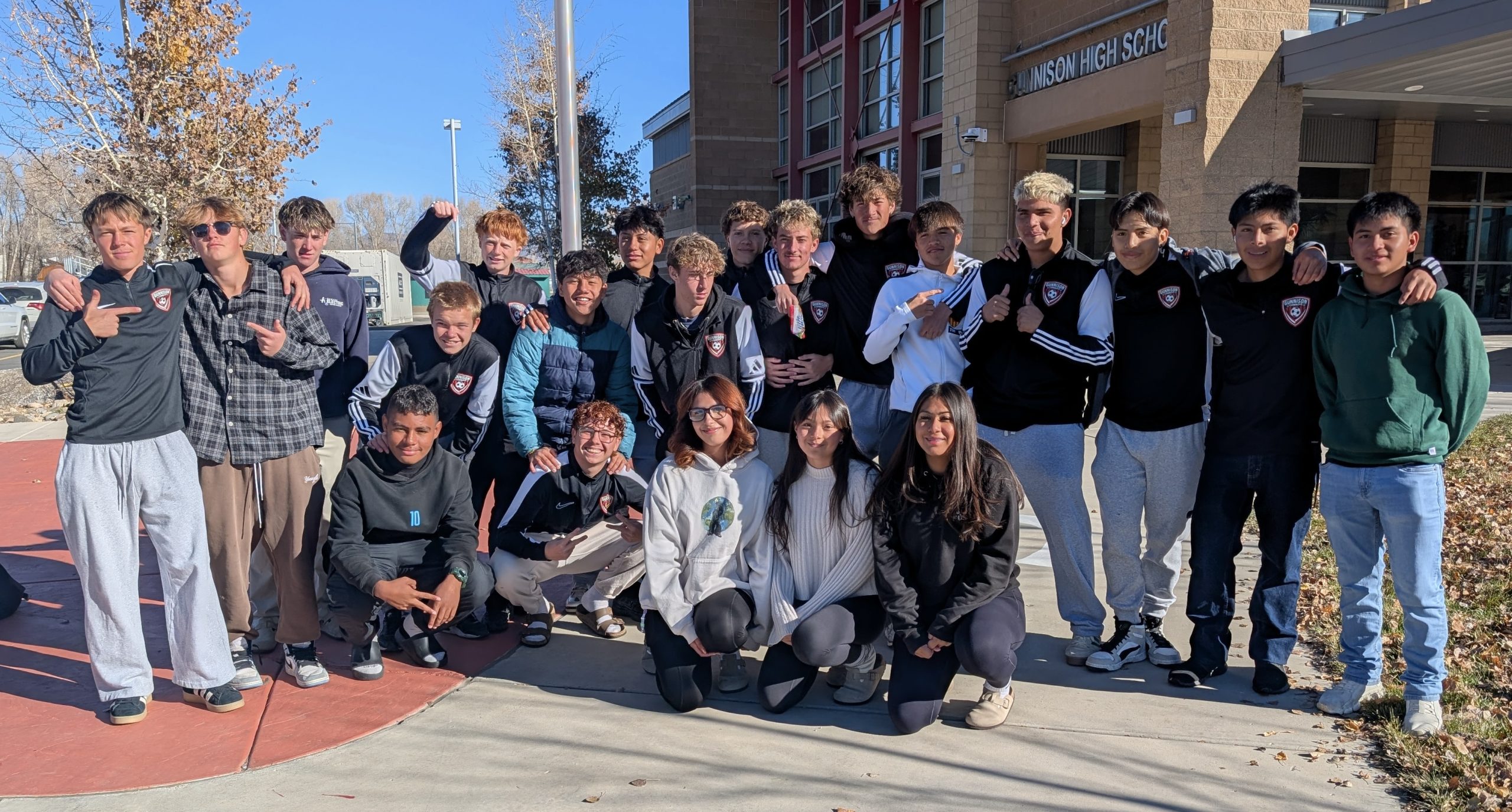 the Fall 2025 Gunnison High School Boys soccer team and managers smile for a group photo in front of the school